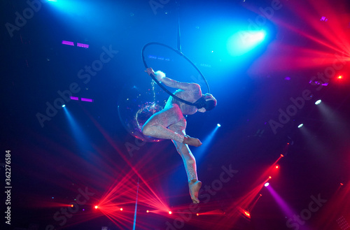 Flexible young woman make performance on aerial hoop, flexible back on aerial hoop, aerial circus show, yellow purple blue red light. Flexible woman gymnast upside down on hoop. Night club performance