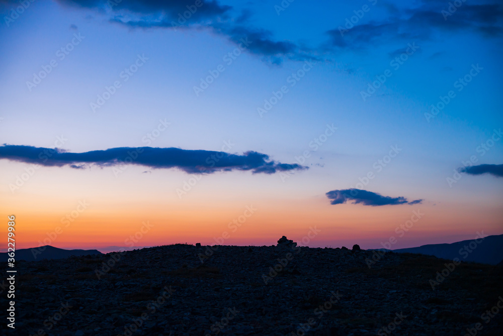Atmospheric dawn landscape with dark silhouette of tour on pass top under vivid orange pink lilac gradient sky with blue clouds on sunset. Beautiful mountain view to stony hill silhouette on sunrise.