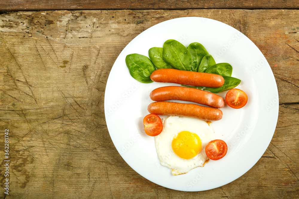 Fried egg with sausages and cherry tomatoes in a white plate on a wooden table. Fried egg with sausages and cherry tomatoes in a white plate on a wooden table.