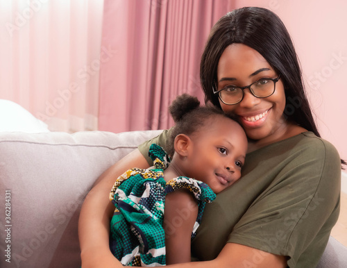 Foto Head shot portrait of happy African American mother cuddles a lovely little daughter sitting on the sofa and looking at camera