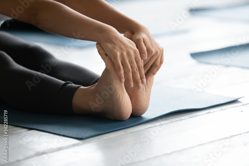 Close up young fit woman sitting on floor mat  holding feet in hands  stretching muscles in seated forward bend position  enjoying morning yoga exercises in modern class studio center indoors.
