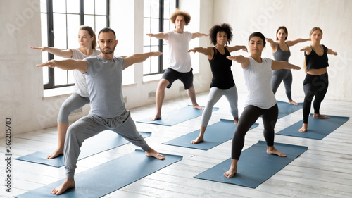 Focused sporty handsome young male trainer leading yoga class for concentrated multiracial students, practicing virabhadrasana second warrior II position together at yoga training in modern studio.
