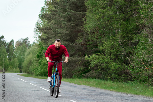 Fototapeta Naklejka Na Ścianę i Meble -  A man in a tracksuit on a bicycle rides on a road in the forest. The concept of a healthy lifestyle, cardio training. Copyspace.