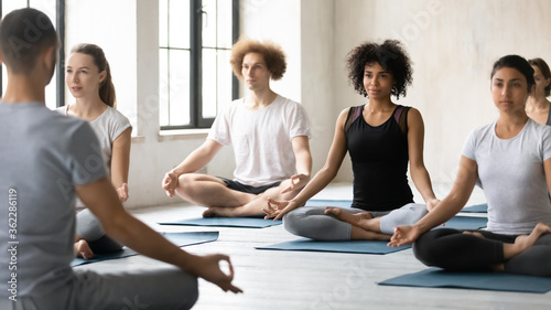 Focused peaceful young mixed race people sitting in open hips padmasana lotus pose, looking at male instructor at yoga master class in modern studio club interior, learning meditating indoors.