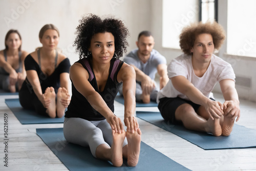 Sporty multiracial young people sitting in rows on floor mats in seated forward bend exercise pose, enjoying morning group yoga class in modern studio, happy diverse beginners working pit together.