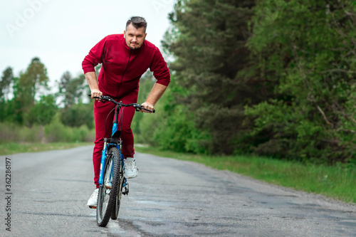 A man in a tracksuit on a bicycle rides on a road in the forest. The concept of a healthy lifestyle, cardio training. Copyspace. © Aliaksandr Marko