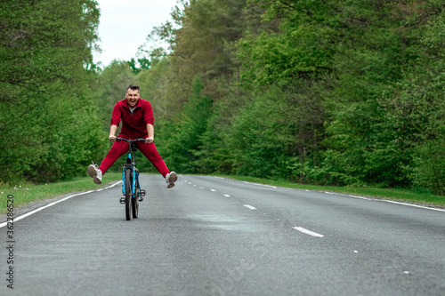 Fototapeta Naklejka Na Ścianę i Meble -  Happy male riding bike with legs up. The road in the forest. The concept of a healthy lifestyle, cardio training. Copyspace.