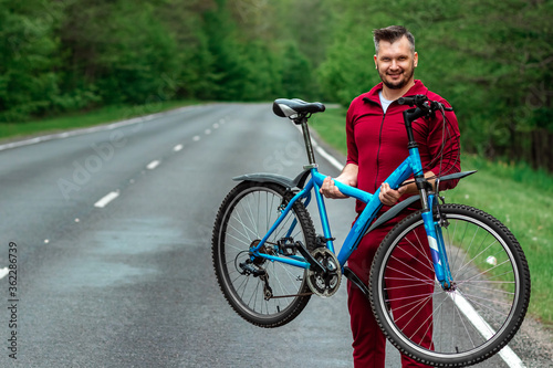 Athletic Man in a tracksuit holds a bicycle on his shoulders while standing on the road in the forest. The concept of a healthy lifestyle, cardio training. Copyspace.