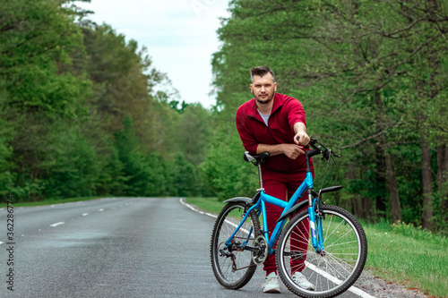 A man in a tracksuit stands next to a bicycle on a road in the forest. The concept of a healthy lifestyle, cardio training. Copyspace. © Aliaksandr Marko
