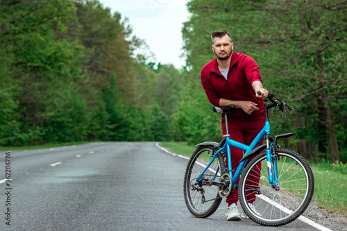 Fototapeta Naklejka Na Ścianę i Meble -  A man in a tracksuit stands next to a bicycle on a road in the forest. The concept of a healthy lifestyle, cardio training. Copyspace.