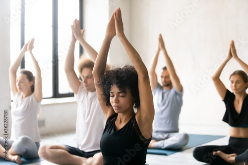 Head shot motivated young peaceful african american woman learner meditating with closed eyes and raised namaste sign arms, enjoying group yoga training with diverse people in modern sport club.