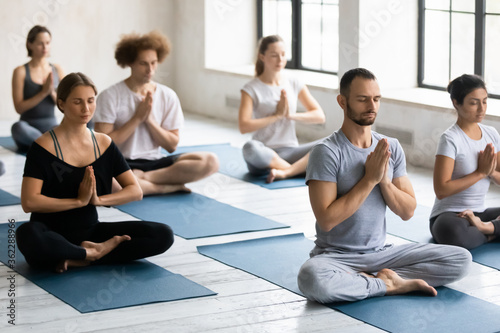 Young motivated multiracial people in activewear sitting on floor mats in lotus position with folded in namaste hands, feeling thankful after workout or enjoying meditating with closed eyes indoors.