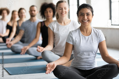 Focus on happy young pretty indian ethnicity woman sitting in padmasana lotus pose on floor mat with diverse multiracial people meditating in row with closed eyes, enjoying yoga class together.