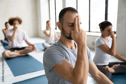 Head shot young mindful male trainer leading yoga group class, showing Alternate Nostril Breathing to motivated diverse students, practicing nadi shodhana pranayama together finishing workout indoors. photo