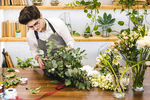 Attractive young male florist with glasses and apron works in a flower shop.
