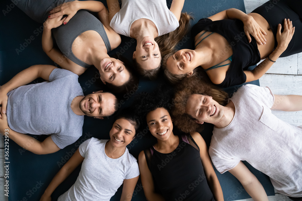 Φωτογραφία Top above view smiling beautiful mixed race people in sportswear lying on floor mats, looking at camera, relaxing after intensive yoga gym workout together, engaged in team building activity indoors