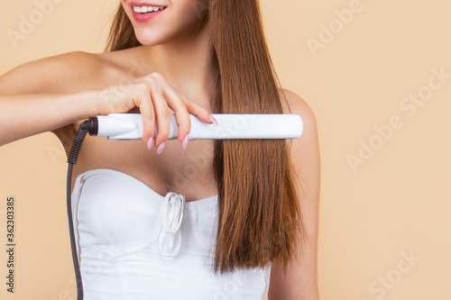Smiling woman ironing long hair with flat iron. Happy young woman straightening hair with straightener. Portrait of young beautiful girl using styler on her shining hair. Hairstyle photo