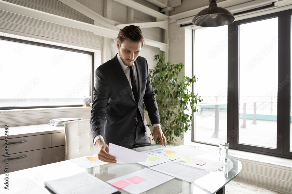 Concentrated Caucasian businessman stand at desk in office manage company financial documents, focused male boss or director work with paperwork at workplace, prepare startup project or report Concentrated Caucasian businessman stand at desk in office manage company financial documents, focused male boss or director work with paperwork at workplace, prepare startup project or report