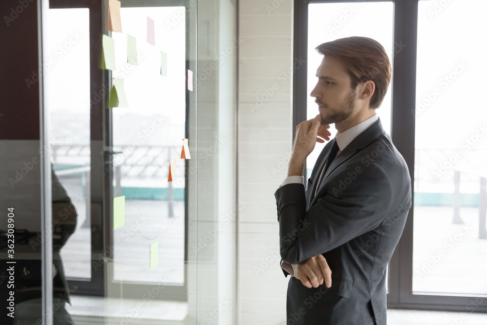 Pensive young Caucasian businessman look at glass wall in office think of project implementation, thoughtful serious male employee brainstorm develop business plan in boardroom, work with sticky notes Pensive young Caucasian businessman look at glass wall in office think of project implementation, thoughtful serious male employee brainstorm develop business plan in boardroom, work with sticky notes