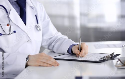 Unknown male doctor sitting and working with clipboard of medication history record in clinic at his working place, close-up. Young physician at work. Perfect medical service, medicine concept
