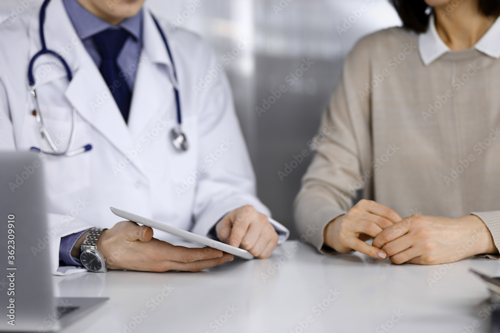 Unknown male doctor and patient woman discussing something while sitting in clinic and using tablet computer. Best medical service in hospital, medicine, pandemic stop