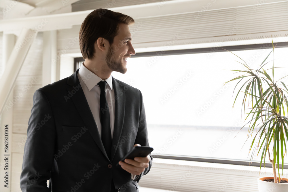 Smiling young businessman hold cellphone look in window distance thinking or visualizing, happy Caucasian male employee use smartphone, ponder plan future career success, business vision concept