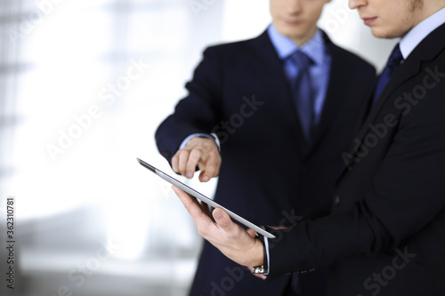 Business people use a tablet computer for discussion of their new project, standing in a modern office. Unknown businessman or male entrepreneur with a colleague at workplace. Teamwork and partnership