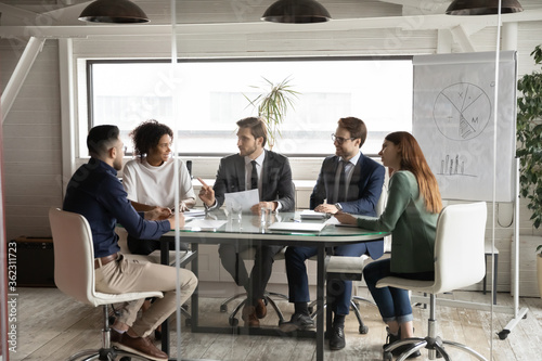 Smiling multiethnic businesspeople sit at desk in office talk discuss business paperwork document together, multiracial employees cooperate at meeting in boardroom, teamwork, collaboration concept