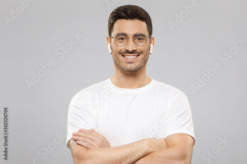 Young handsome man wearing white t-shirt, round glasses and wireless earphones, standing with crossed arms, smiling confidently, isolated on gray background