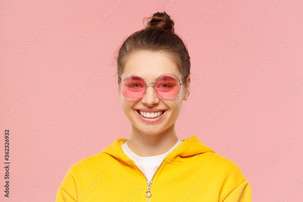 Close up portrait of young smiling girl wearing yellow casual hoodie and round colored glasses, isolated on pink background