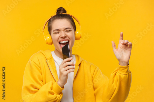 Young girl in yellow hoodie and wireless headphones, singing with closed eyes, holding smartphone as microphone, having fun, isolated on studio background