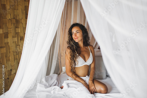 Beautiful tanned young woman awakening in white bed. Happy wake up and start new day. Leisure and rest. Wellbeing and carefree concept. Long curly hair. Stay home. © Oleg Breslavtsev