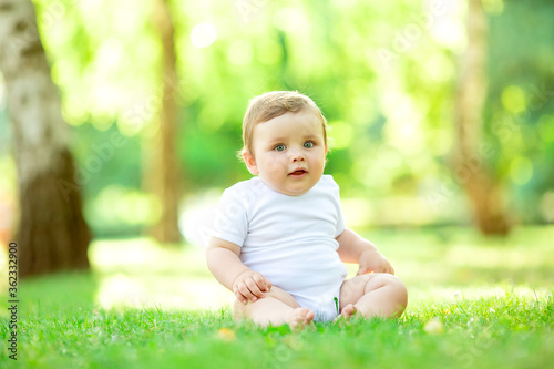 Cute toddler boy of 10 months in white bodysuit sits on green summer grass, space for text