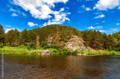 River in summer on the background of rocks, pines, blue sky and white clouds