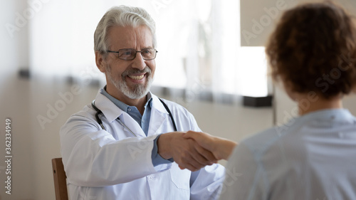 Smiling mature Caucasian male doctor in white medical uniform shake hand get acquainted greeting with patient, happy senior man GP handshake client at consultation meeting, close healthcare insurance