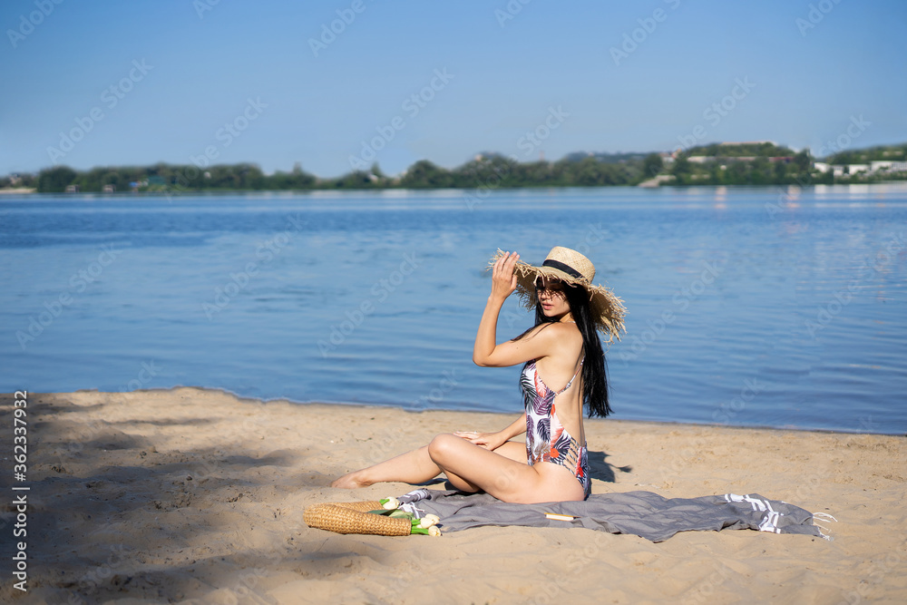 Naklejka premium dark-haired beautiful girl in a swimsuit sitting on the beach in a straw hat 