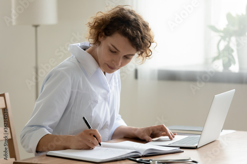 Young Caucasian female doctor in white medical uniform sit at desk work on laptop make note in register, woman nurse or GP fill patient anamnesis in journal, use computer in private clinic