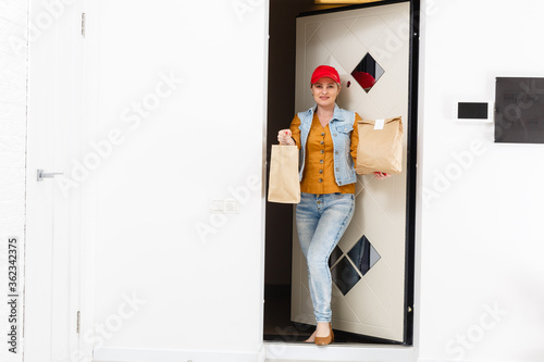 Woman in red cap giving fast food order home background. Female courier holding paper packet with food. Products delivery from shop or restaurant to home. Copy space