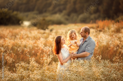 Mom, dad and their little daughter are having fun in a chamomile field near the forest. They are hugging and laughing. Image with selective focus.