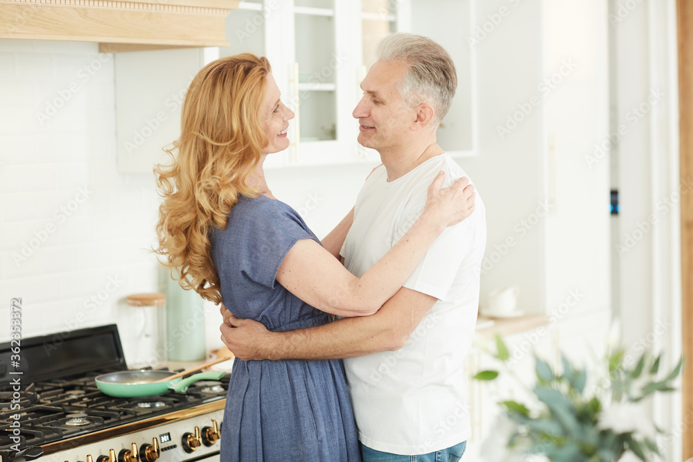 Waist up portrait of smiling mature couple embracing and looking at each other while standing in white kitchen interior at home, copy space Waist up portrait of smiling mature couple embracing and looking at each other while standing in white kitchen interior at home, copy space