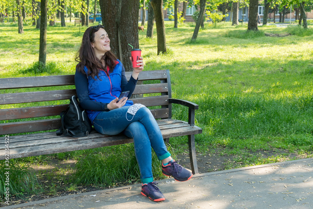Obraz premium Beautiful Caucasian woman with long brown hair sitting in the park wearing a protective mask drinking a cup of tea or coffee and smiling. toned