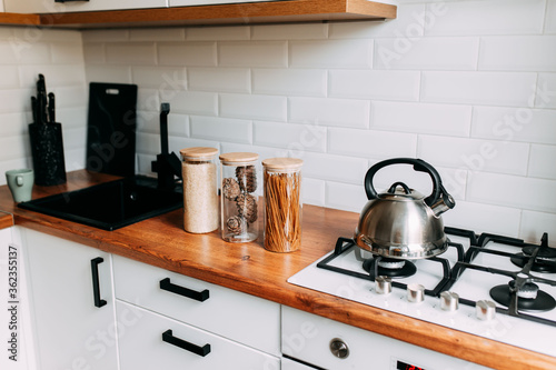 Glass jars in kitchen. Metallic teapot on the gas oven. Bright kitchen interior. White modern dining room. Wooden complete kitchen with gas oven.