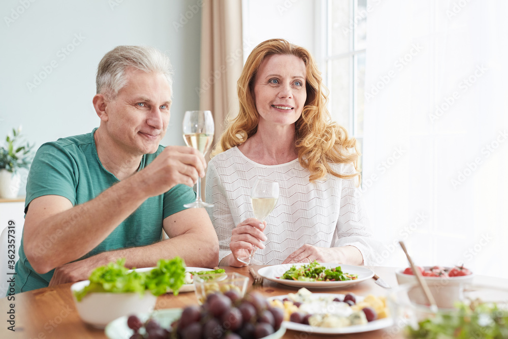 Portrait of loving mature couple raising champagne glasses while enjoying romantic dinner at home, copy space