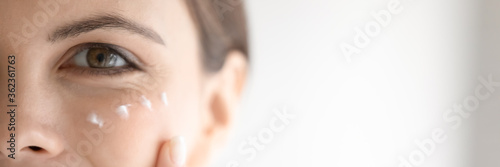 Close up wide cropped image female eye and cheekbone with white face cream, beautiful young woman applying moisturizing lotion, looking at camera, enjoying skincare routine and spa procedure