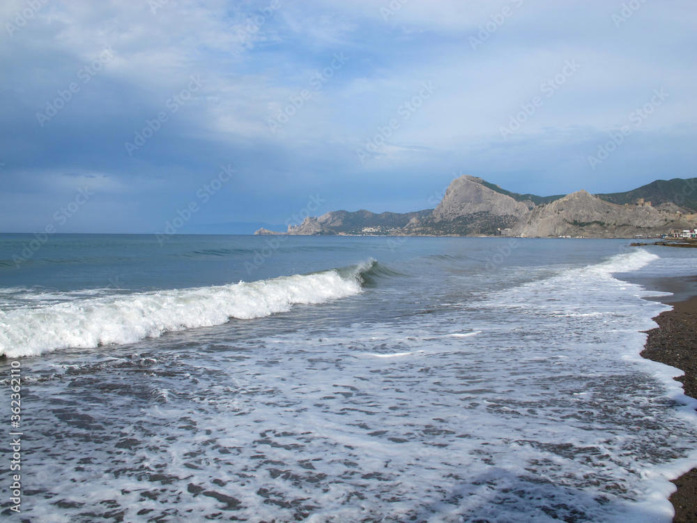 seascape with views of coastal mountains, rocks, the sea with waves, sea foam paints a pattern on the sand on a summer sunny day, Sudak, Crimea