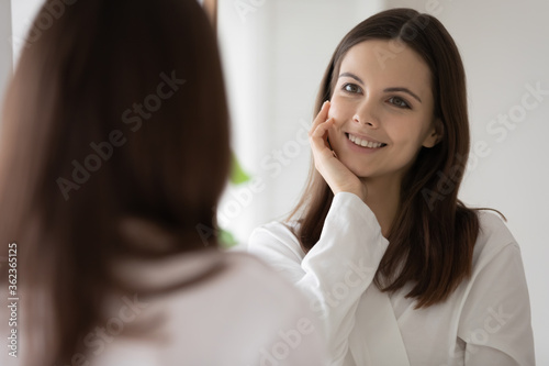 Smiling beautiful young woman looking at mirror in bathroom, attractive girl wearing white bathrobe touching perfect healthy smooth skin, enjoying morning routine, everyday skincare procedure