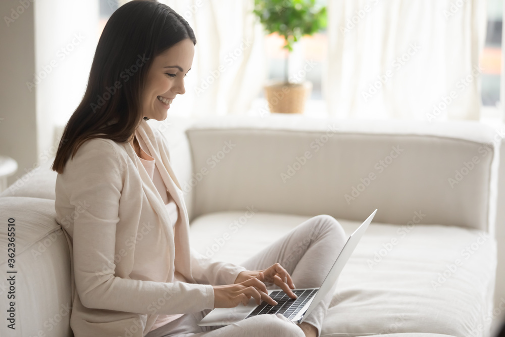 Smiling overjoyed young woman sitting on cozy couch at home, using laptop, happy girl looking at computer screen, chatting in social network, having fun with gadget, freelancer working online Smiling overjoyed young woman sitting on cozy couch at home, using laptop, happy girl looking at computer screen, chatting in social network, having fun with gadget, freelancer working online