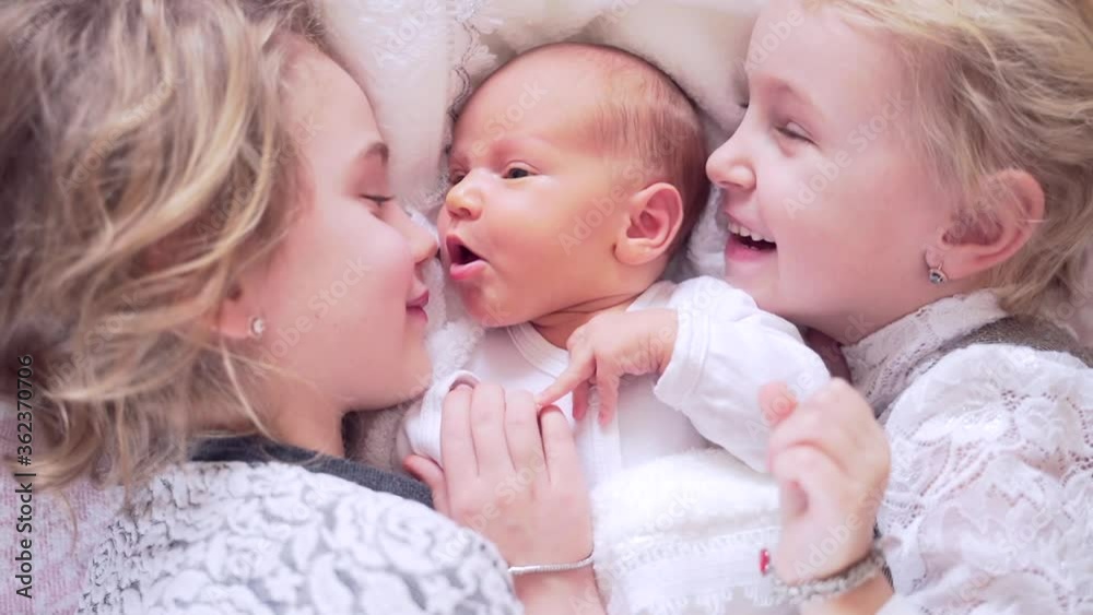 Three cute little children are lying on the bed. The sisters hug and kiss the newborn baby. Portrait of girls lying happy hugging close up. Happy and smiling together Three cute little children are lying on the bed. The sisters hug and kiss the newborn baby. Portrait of girls lying happy hugging close up. Happy and smiling together