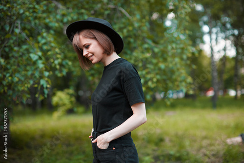 Portraif of a young cute modern millennial hipster girl in a hat dressed in black is relaxing in a city Park.