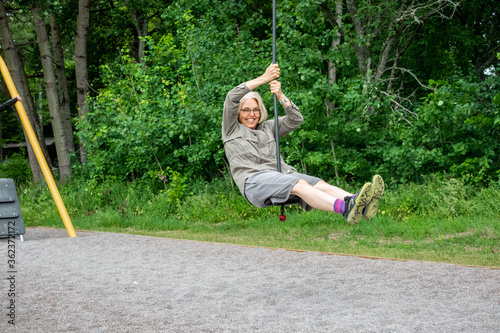 Mature woman with glasses riding a zip-line at an outdoor playground at a public park.
